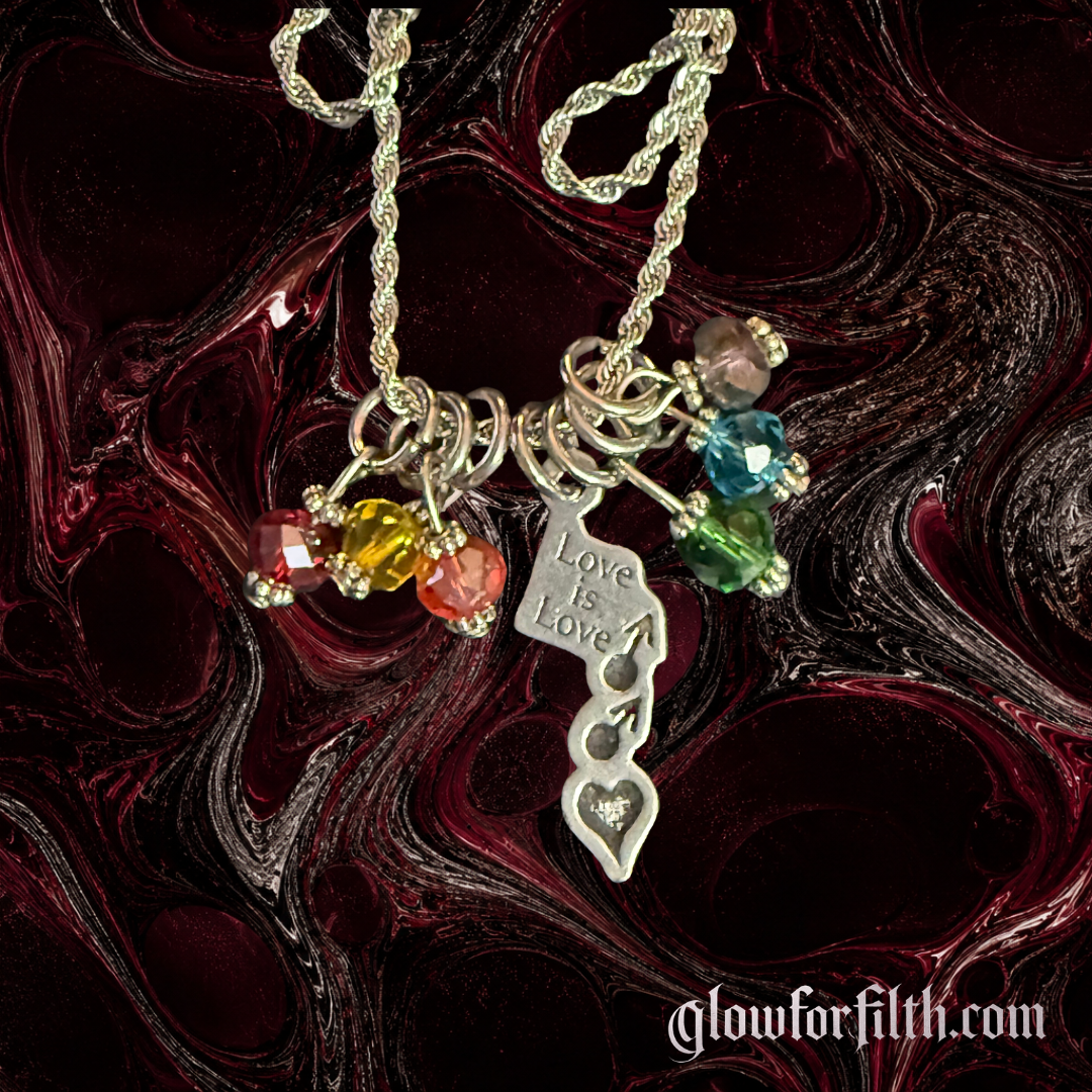 Silver-tone rope chain necklace featuring a lead-free pewter Love Is Love pendant with male-male symbols and rainbow crystal accents displayed on a gothic burgundy and black marbled background Deatiled Shot.