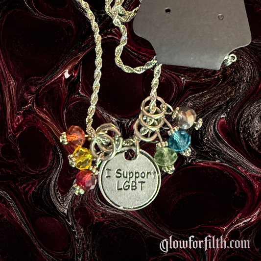 American pewter I Support LGBT necklace with six rainbow crystal accent beads on a stainless steel rope chain against a dark red and black background.