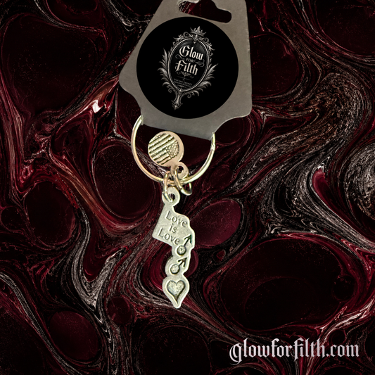 Silver-tone keychain featuring a Love Is Love charm with interconnected male symbols displayed on a gothic burgundy and black marbled background.