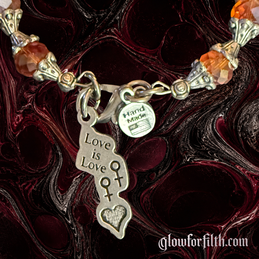 Handmade crystal bracelet featuring a lead-free pewter Love Is Love charm with female-female symbols and faceted crystal beads displayed on a gothic burgundy and black marbled background detailed shot.