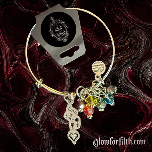 Silver-tone expandable bangle bracelet featuring a lead-free pewter Love Is Love charm with female-female symbols and rainbow crystal accents displayed on a gothic burgundy and black marbled background.