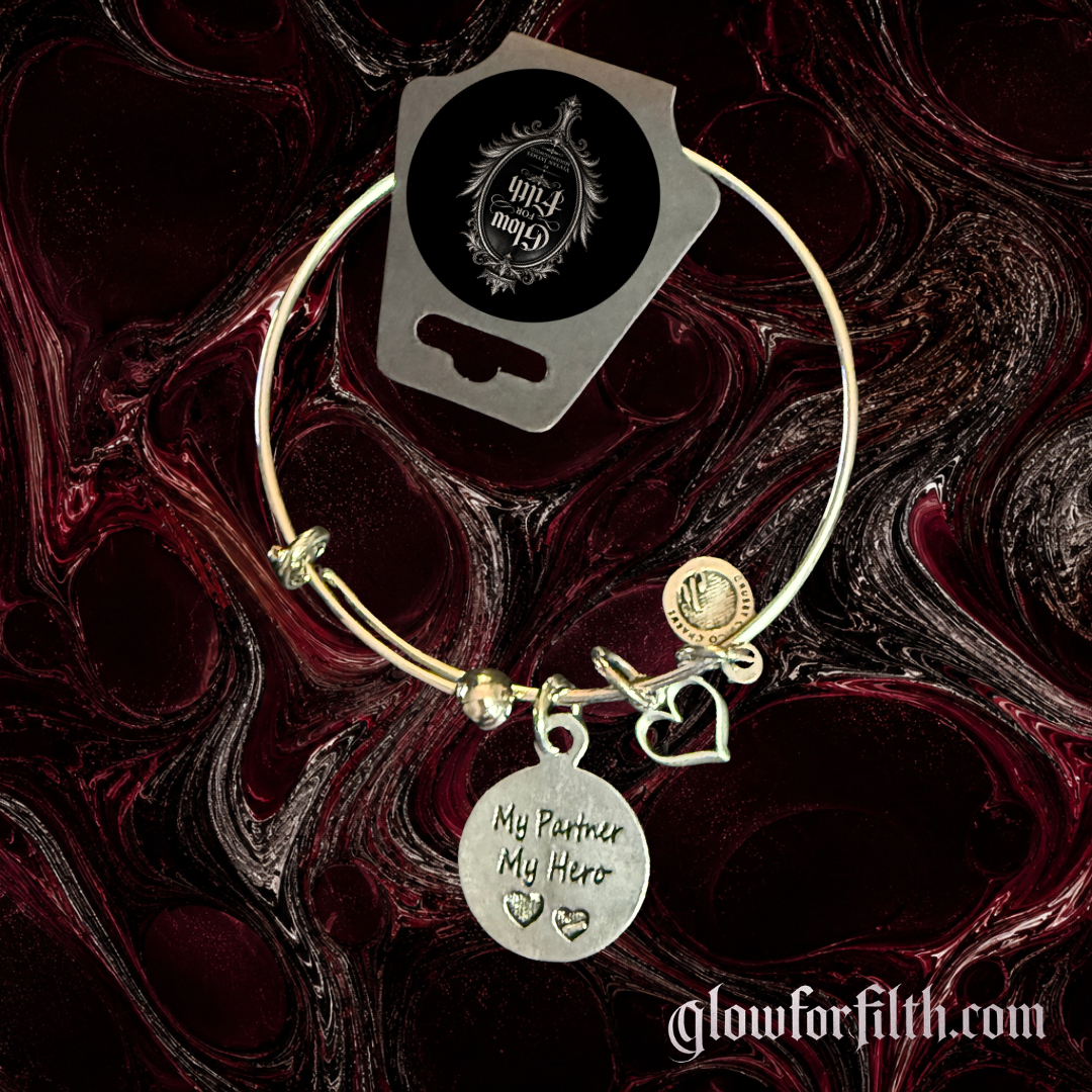Silver-tone expandable bangle bracelet featuring a lead-free pewter My Partner My Hero charm with heart accents displayed on a gothic burgundy and black marbled background.