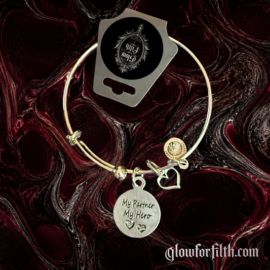 Silver-tone expandable bangle bracelet featuring a lead-free pewter My Partner My Hero charm with heart accents displayed on a gothic burgundy and black marbled background.
