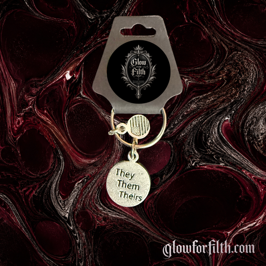 Silver-tone keychain featuring a lead-free pewter They Them Theirs pronoun charm displayed against a burgundy and black gothic marbled background.