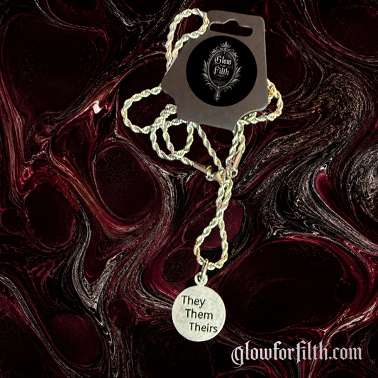 Silver-tone rope chain necklace featuring a lead-free pewter They Them Theirs pronoun charm displayed on a gothic burgundy and black marbled background.